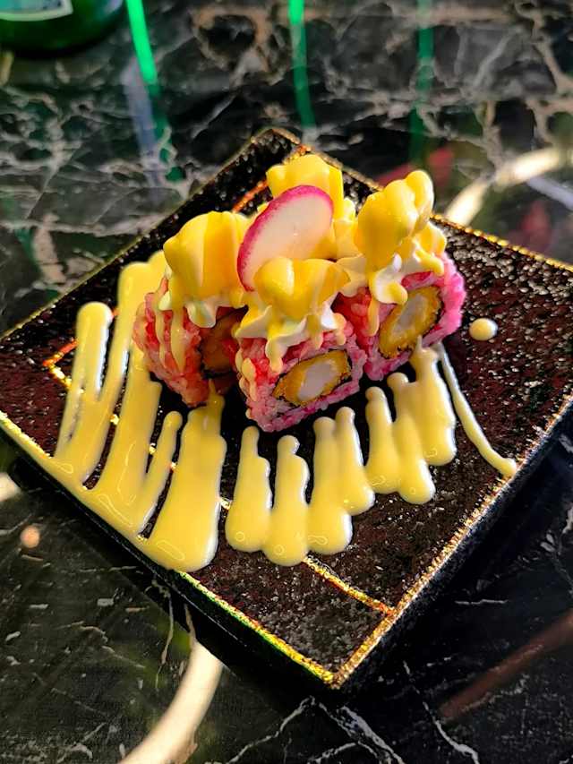 Plat de sushi coloré avec plusieurs rouleaux garnis de tranches de mangue et d'une sauce crémeuse, le tout sur un extérieur texturé rose. Des tranches de radis ajoutent une touche de couleur, le tout présenté de manière artistique sur une assiette sombre et brillante.